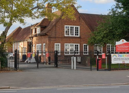 A traditional brick building stands behind a black iron fence, surrounded by trees and a clear sky.