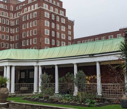 Exterior of a building with a covered area containing columns, large brick building in the background with a lot of white timber sash windows