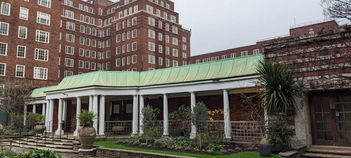 Exterior of a building with a covered area containing columns, large brick building in the background with a lot of white timber sash windows