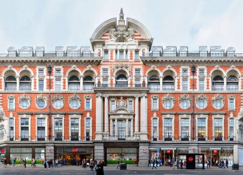 An ornate red and white building with several stories, adorned with sculptures and arches, stands prominently above bustling street-level shops and pedestrians.