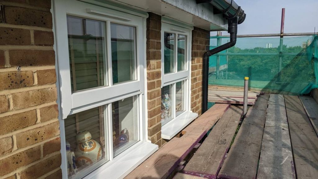 Two white-framed windows on a brick wall, each with a ceramic figurine on the sill, overlooking a wooden deck with greenery in the distance.