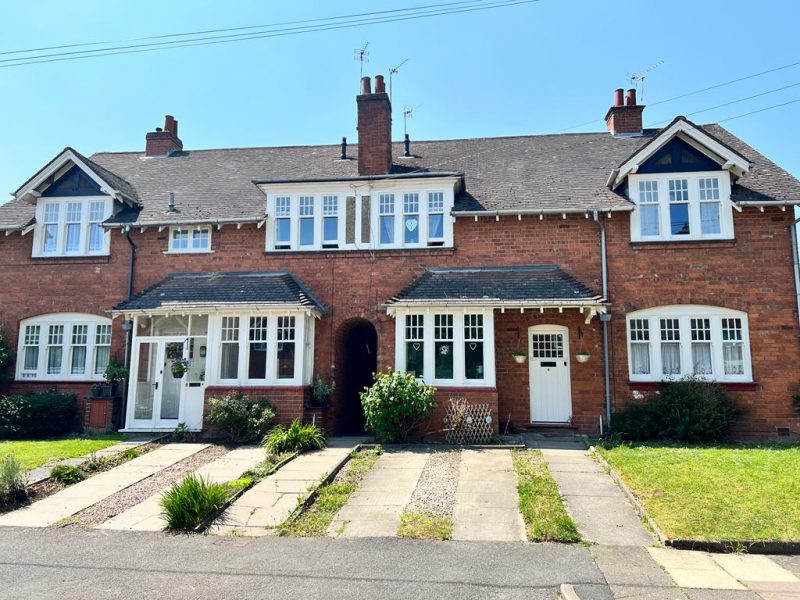 A brick semi-detached house stands under a clear sky, with identical doors and windows, framed by small gardens and a shared central pathway.