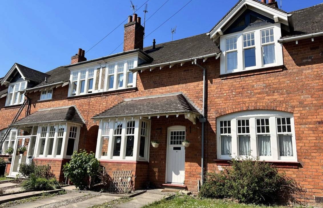 Red-brick terraced houses, with white windows and doors, stand under a clear blue sky, each featuring a small front garden with different plants.