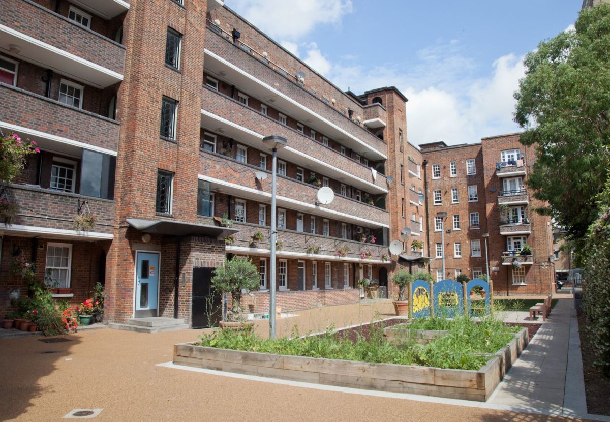 A residential housing estate with a brick facade, multiple balconies, and white-framed windows. The foreground features a courtyard with raised garden beds, plants, and a children’s play area. Some balconies have flower pots, and satellite dishes are mounted on the walls.