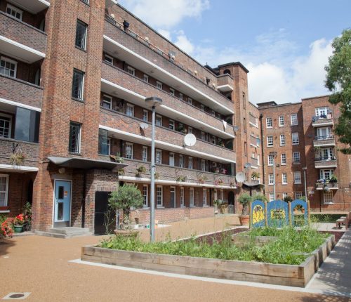 A residential housing estate with a brick facade, multiple balconies, and white-framed windows. The foreground features a courtyard with raised garden beds, plants, and a children’s play area. Some balconies have flower pots, and satellite dishes are mounted on the walls.