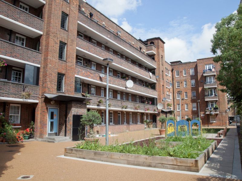 A residential housing estate with a brick facade, multiple balconies, and white-framed windows. The foreground features a courtyard with raised garden beds, plants, and a children’s play area. Some balconies have flower pots, and satellite dishes are mounted on the walls.