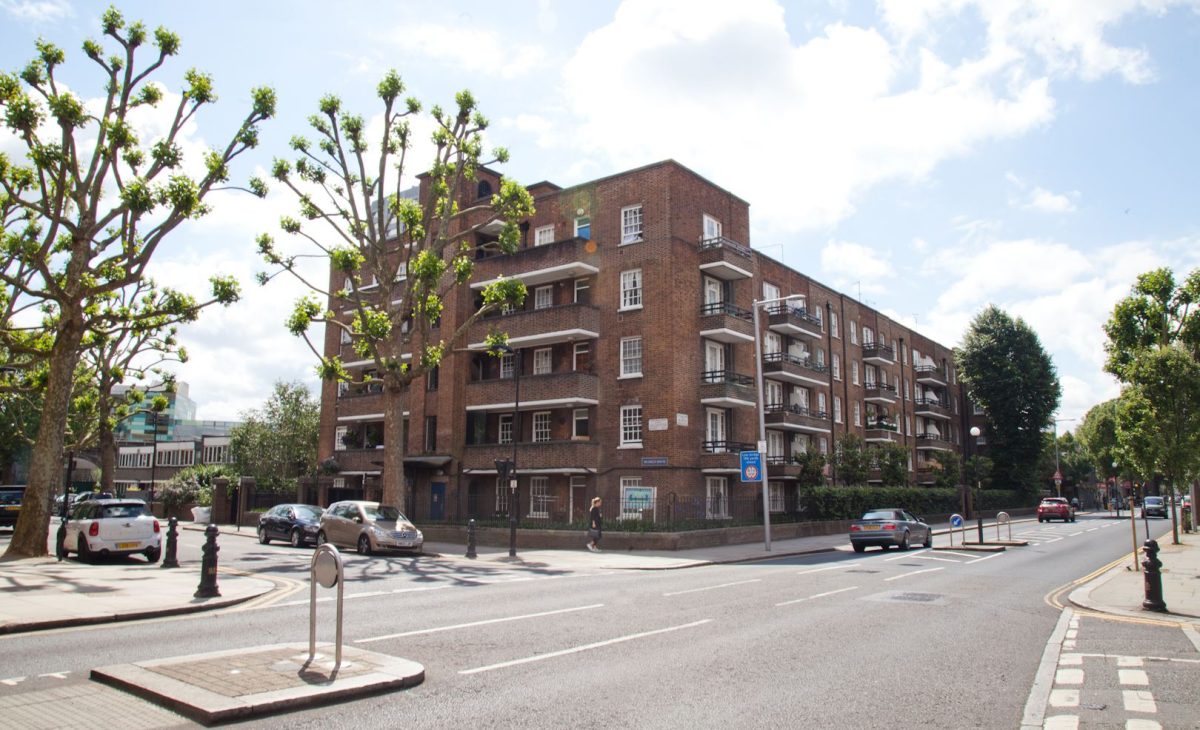 A street view of a brick-built apartment block with multiple balconies, surrounded by a well-maintained urban environment. The road in front has cars parked along the side, a pedestrian crossing, and trees with trimmed branches lining the sidewalk.