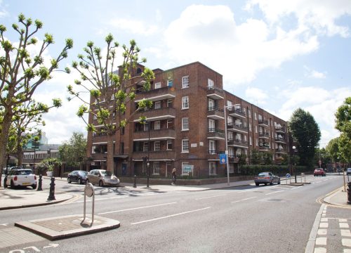 A street view of a brick-built apartment block with multiple balconies, surrounded by a well-maintained urban environment. The road in front has cars parked along the side, a pedestrian crossing, and trees with trimmed branches lining the sidewalk.