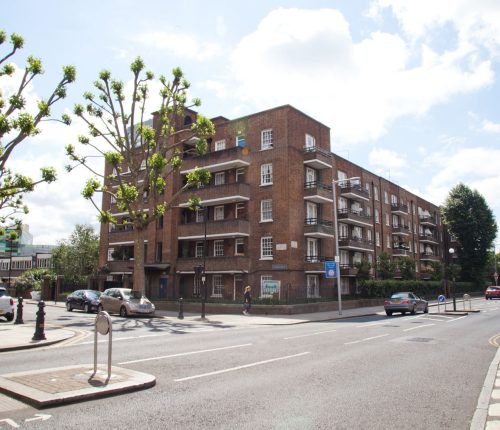 A street view of a brick-built apartment block with multiple balconies, surrounded by a well-maintained urban environment. The road in front has cars parked along the side, a pedestrian crossing, and trees with trimmed branches lining the sidewalk.