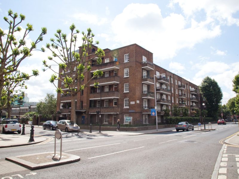 A street view of a brick-built apartment block with multiple balconies, surrounded by a well-maintained urban environment. The road in front has cars parked along the side, a pedestrian crossing, and trees with trimmed branches lining the sidewalk.